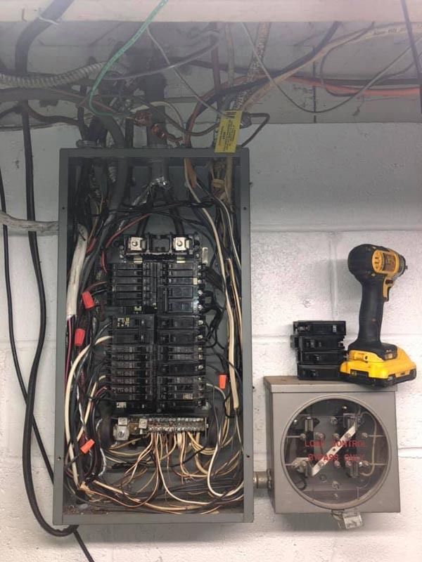 Electrical panel with exposed wires, circuit breakers, and meter box against a white brick wall. A cordless drill rests nearby.
