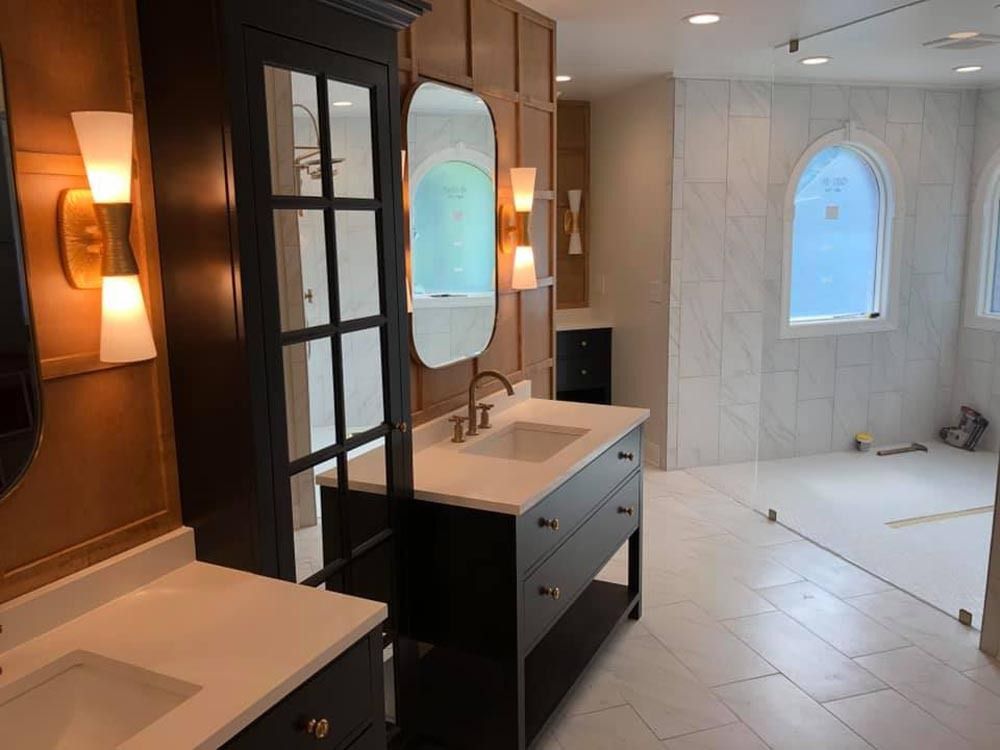 Bathroom with two sinks, dark cabinets, gold sconces, and a large arched window with white tile.
