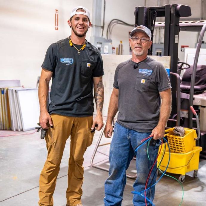 Two men in work clothes, smiling in a warehouse setting, holding tools and wires.