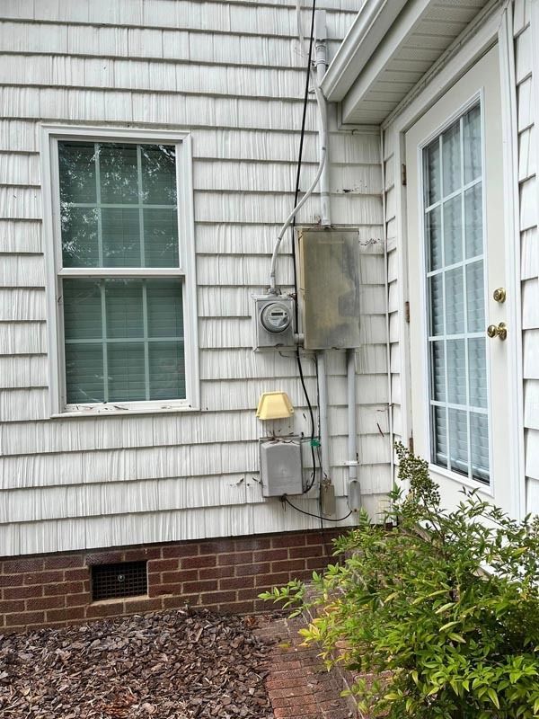 Exterior of a house with electrical boxes and a window and door. White siding, brick foundation, and a bush.