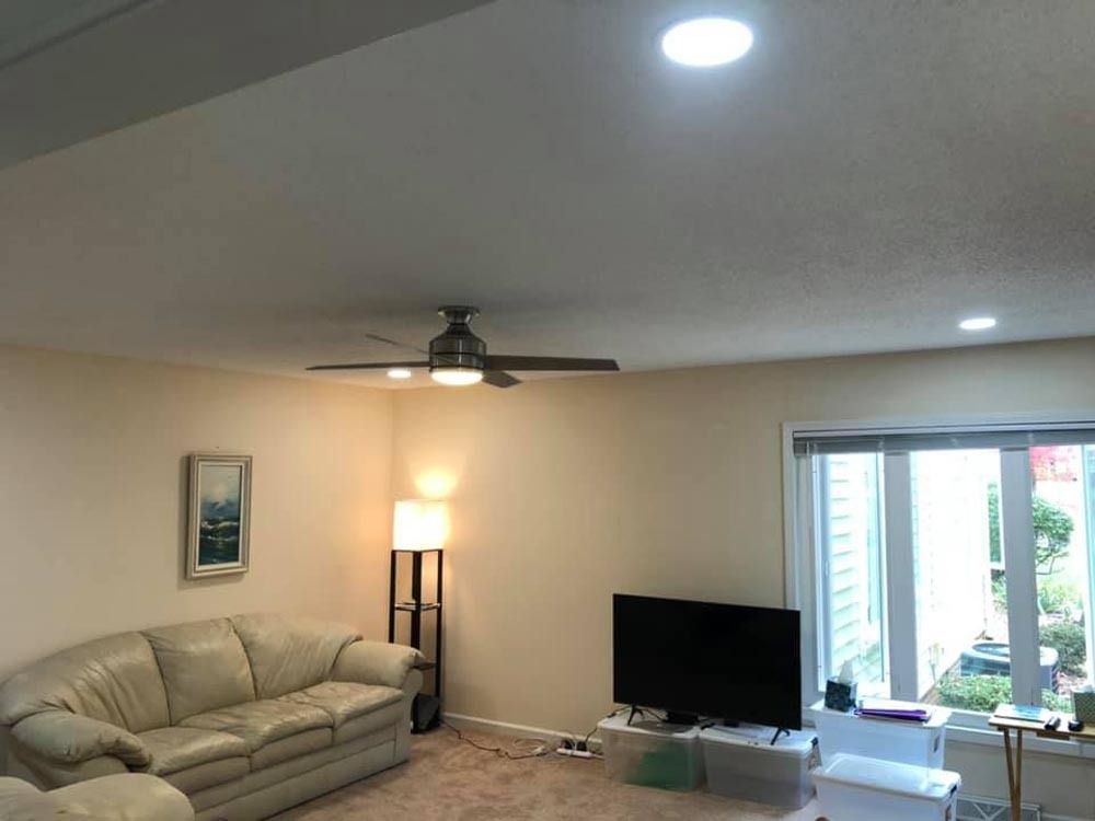 Living room with beige walls, sofa, TV, ceiling fan, and large window.
