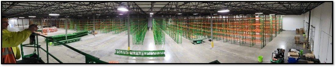 Warehouse interior with rows of storage shelves, green and orange, concrete floor, overhead lighting.