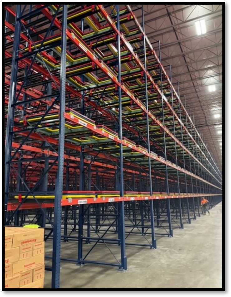 Warehouse with tall, multi-level storage racks; red, yellow, black beams and blue supports; concrete floor.