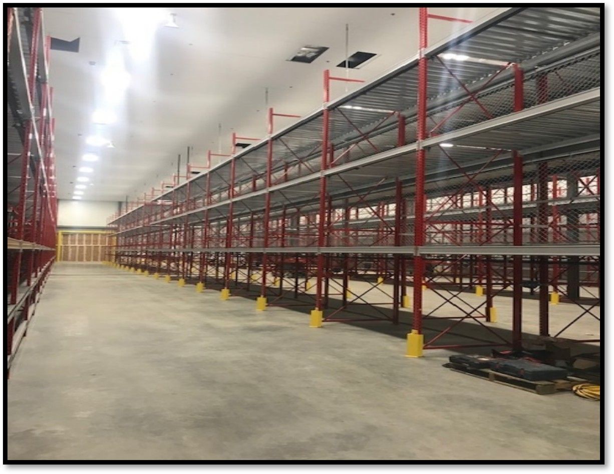 Empty warehouse interior with rows of red shelving, concrete floor, and bright overhead lights.