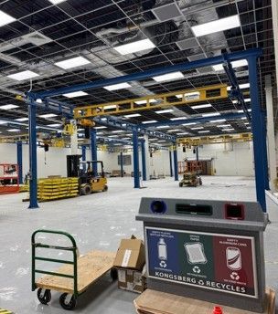 Factory interior with blue and yellow overhead crane system, recycling bins, and a forklift.