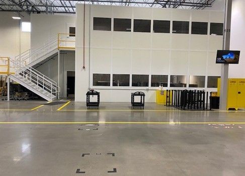 A large industrial interior with a two-story white structure, windows, stairs, and carts. Gray concrete floor with yellow lines.