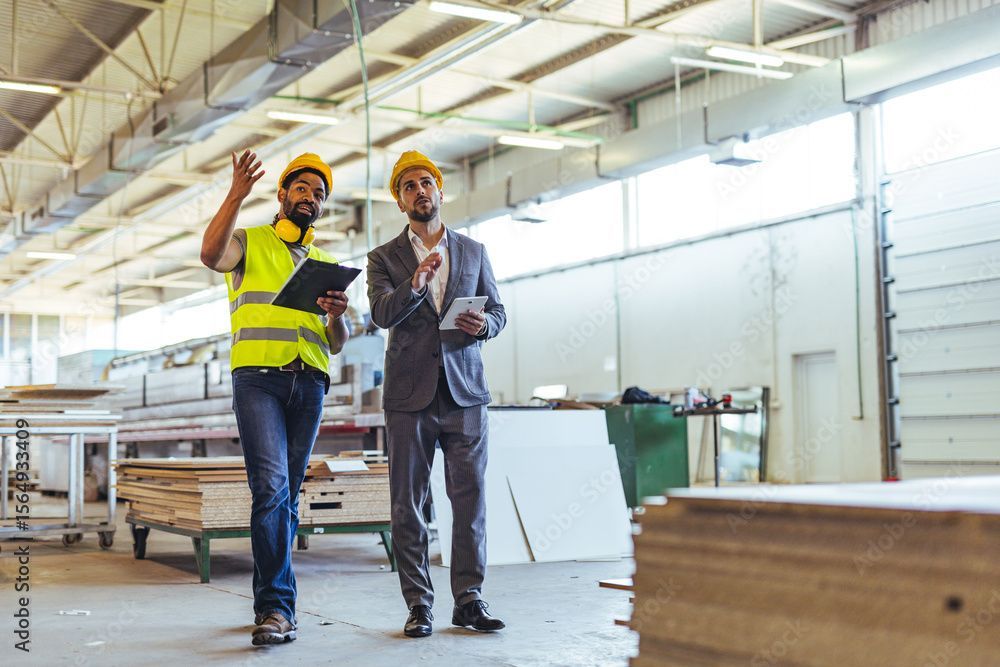 Warehouse worker in a yellow safety vest and hard hat inspects forklift, holding a clipboard, with shelves in background.