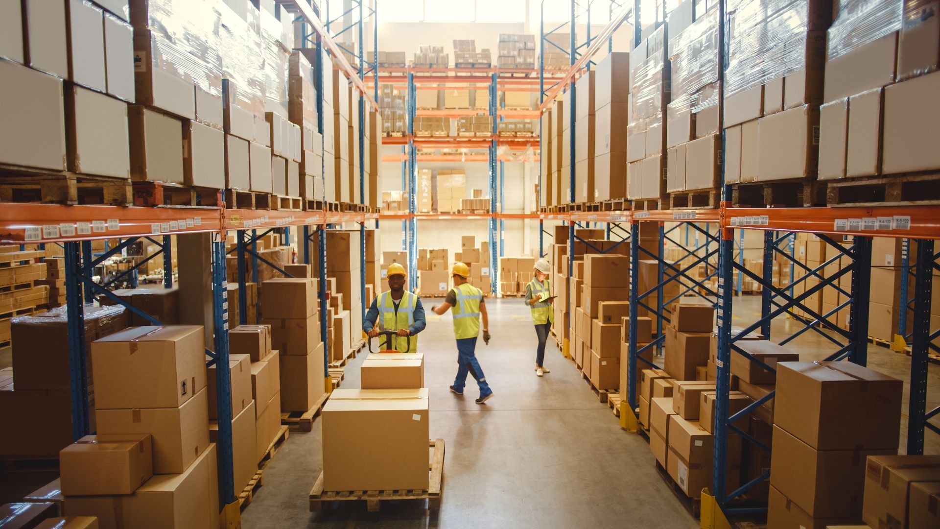 Warehouse interior with workers, stacked boxes on shelving, and pallet jacks.
