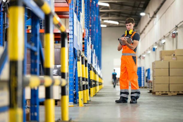 Warehouse worker in orange jumpsuit taking notes near storage racks.