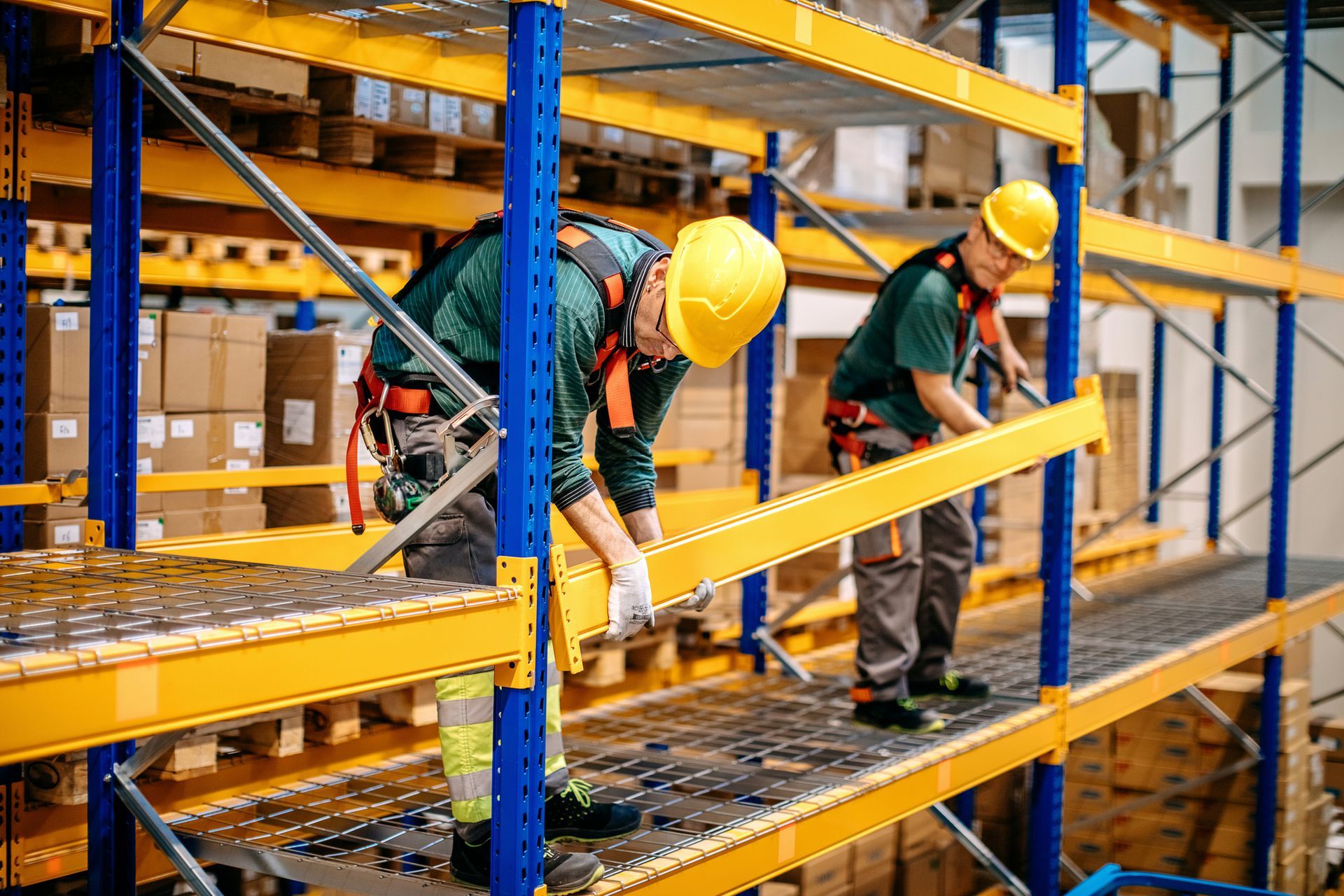 Two workers in hard hats install shelving in a warehouse. They are wearing safety harnesses and gloves. Yellow and blue.