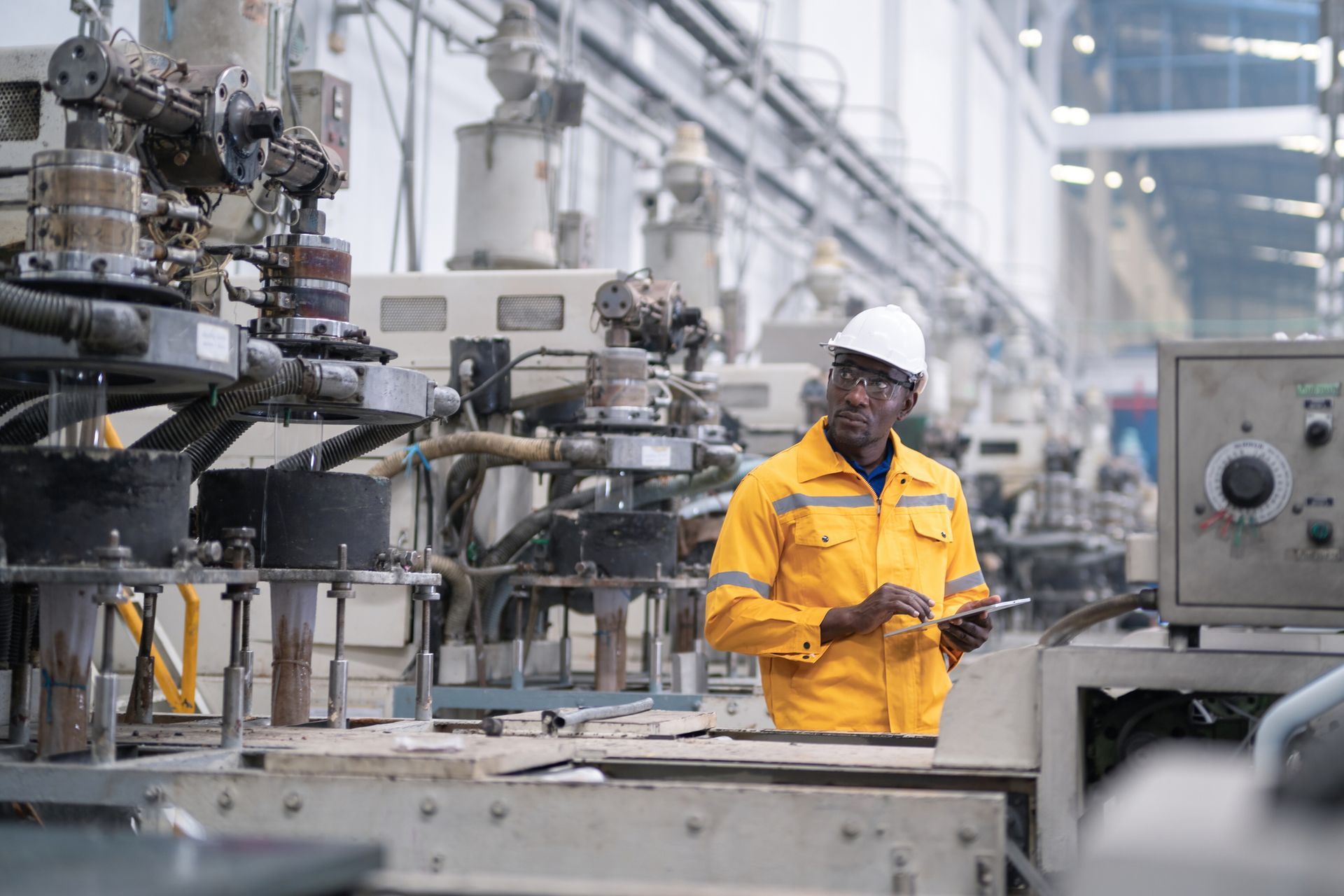 Factory worker in yellow uniform and hard hat using a tablet amidst machinery.