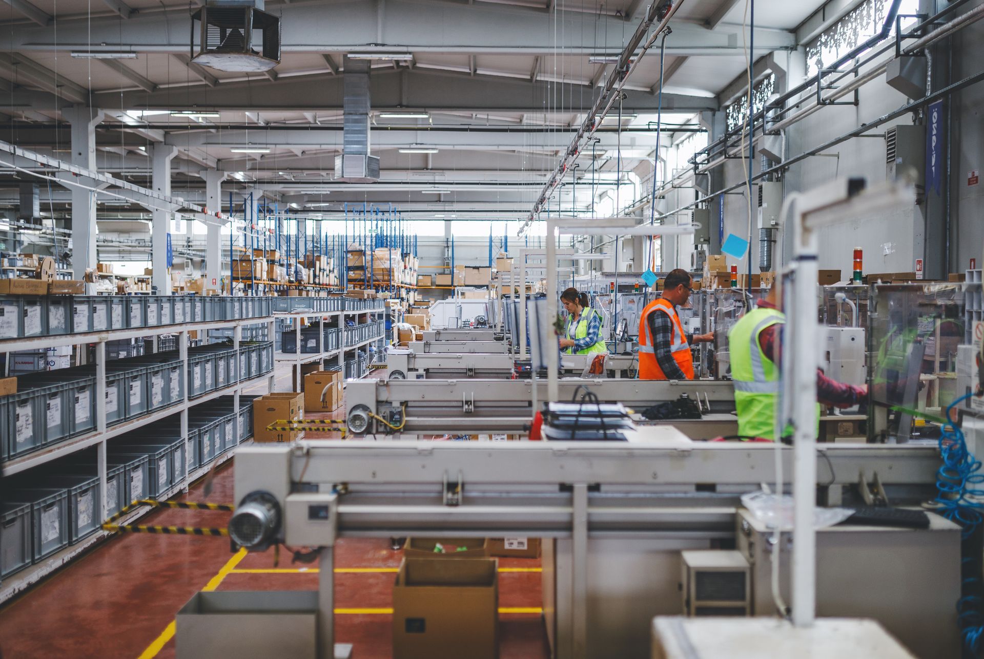Workers at workstations in a factory setting, with shelving, machinery, and safety vests.