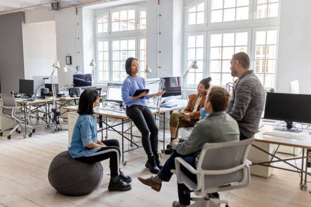 Group of people in an office, gathered for a meeting. One woman holds a tablet, speaking.