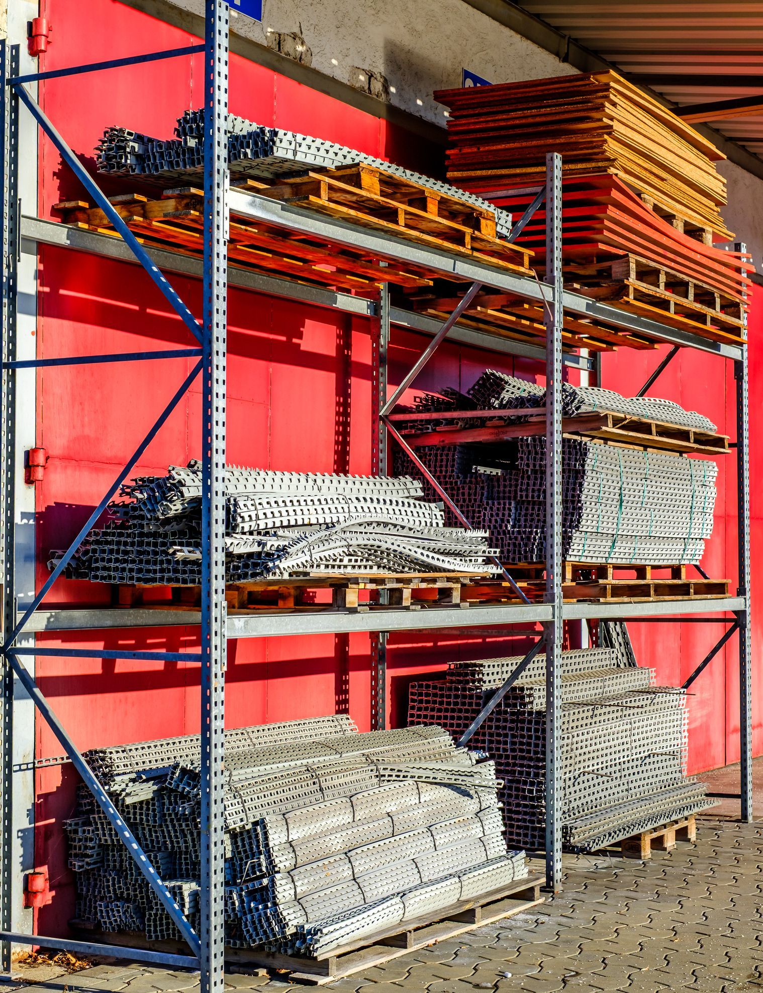 Metal shelving unit filled with pallets and stacked materials against a red wall.