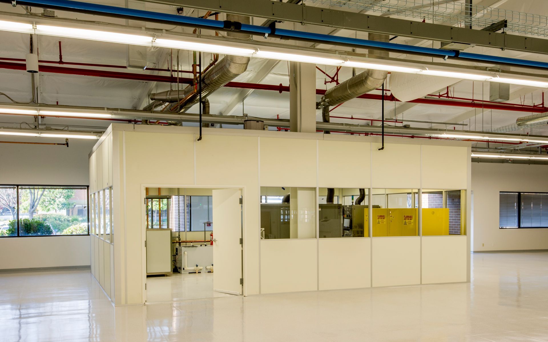 White walled room inside a large industrial space with windows and machinery. Fluorescent lights on the ceiling.