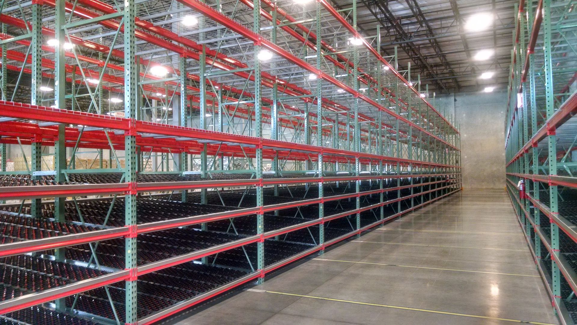 Warehouse interior with rows of empty red and green storage racks. Concrete floor, overhead lighting. Warehouse interior with rows of empty red and green storage racks. Concrete floor, overhead lighting.