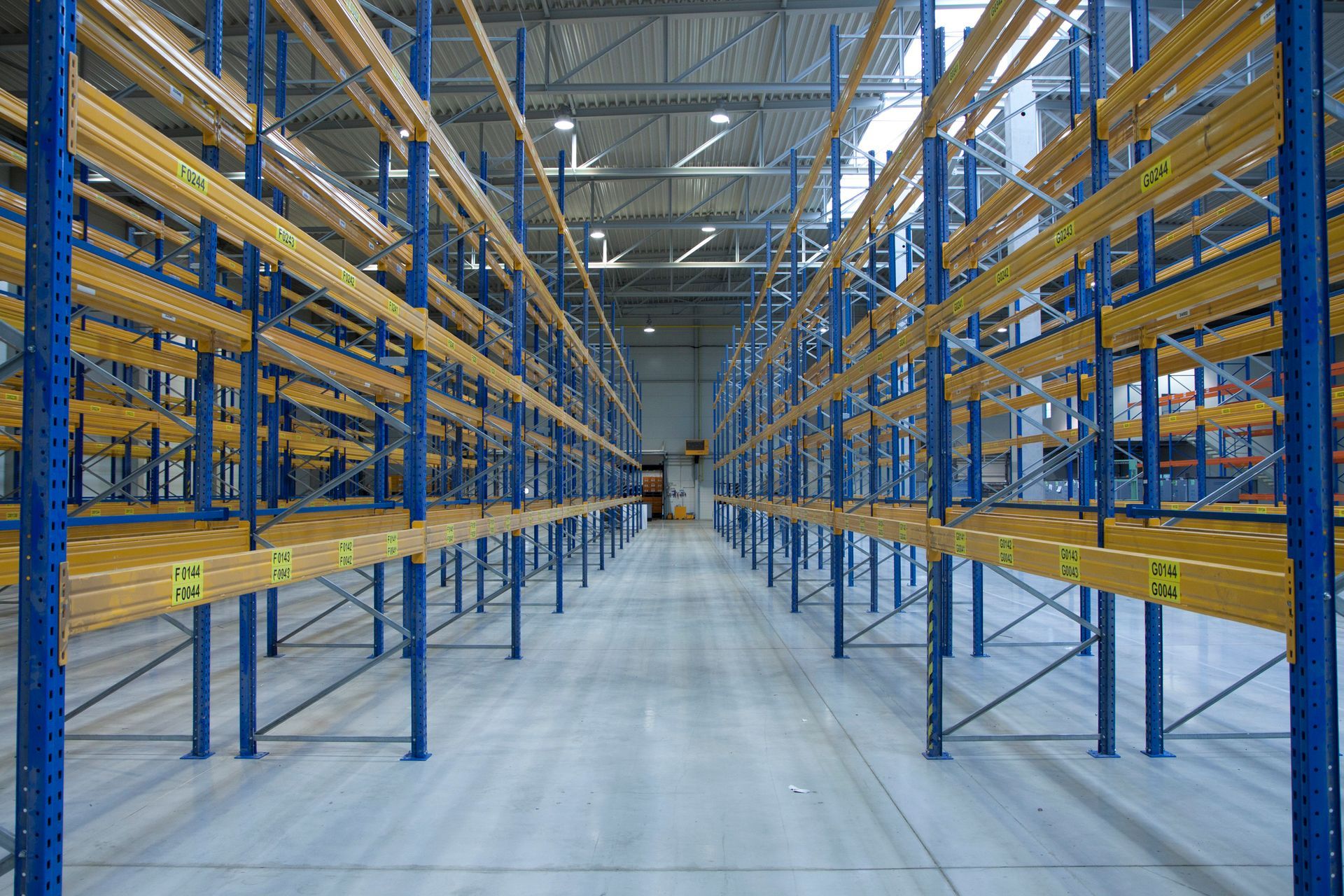 Empty warehouse with rows of tall, blue and yellow shelving units extending toward the back.