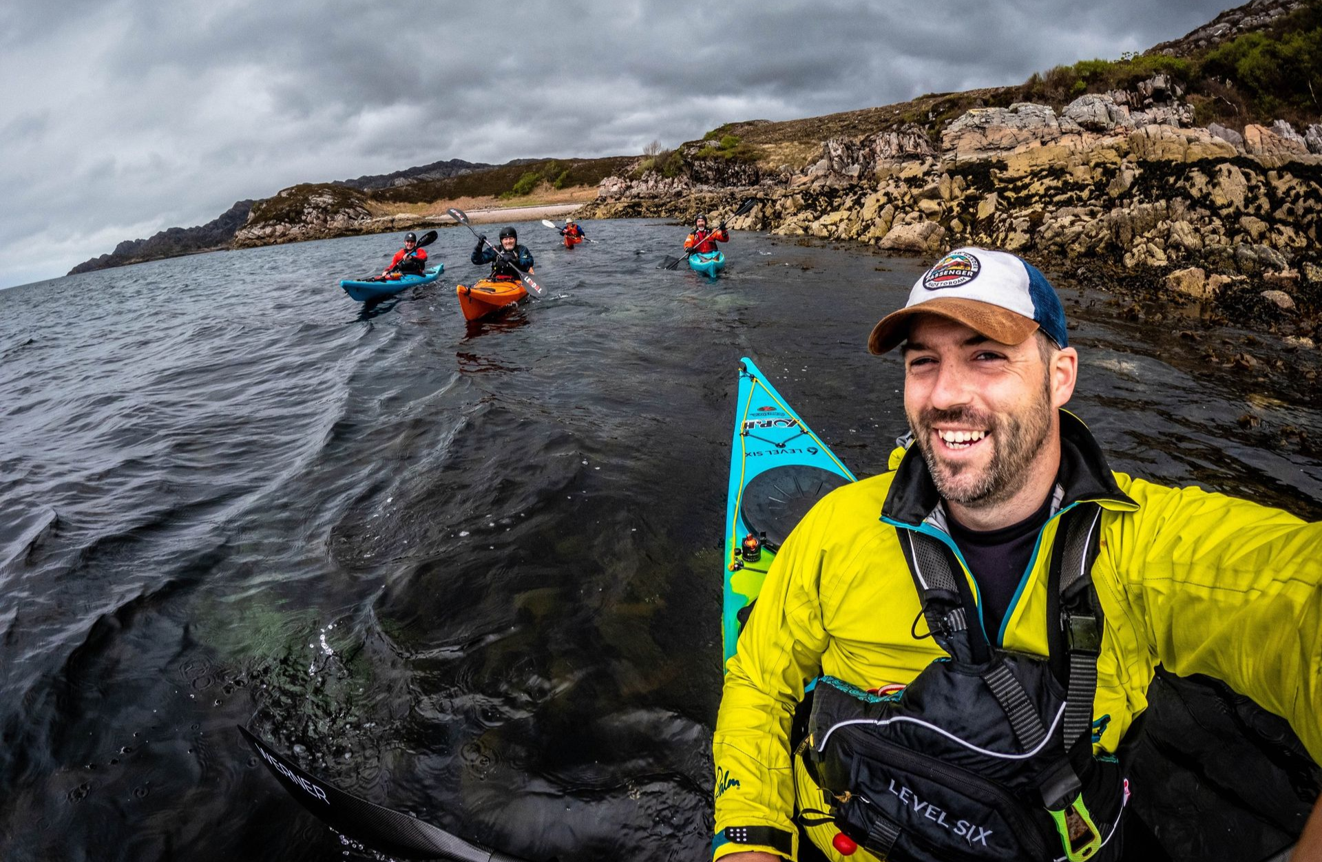 Man in a red jacket kayaking in clear water near a rocky shoreline; smiling.