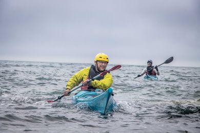 Two people kayaking in choppy, gray water. The person closest smiles, wearing yellow jacket and helmet.