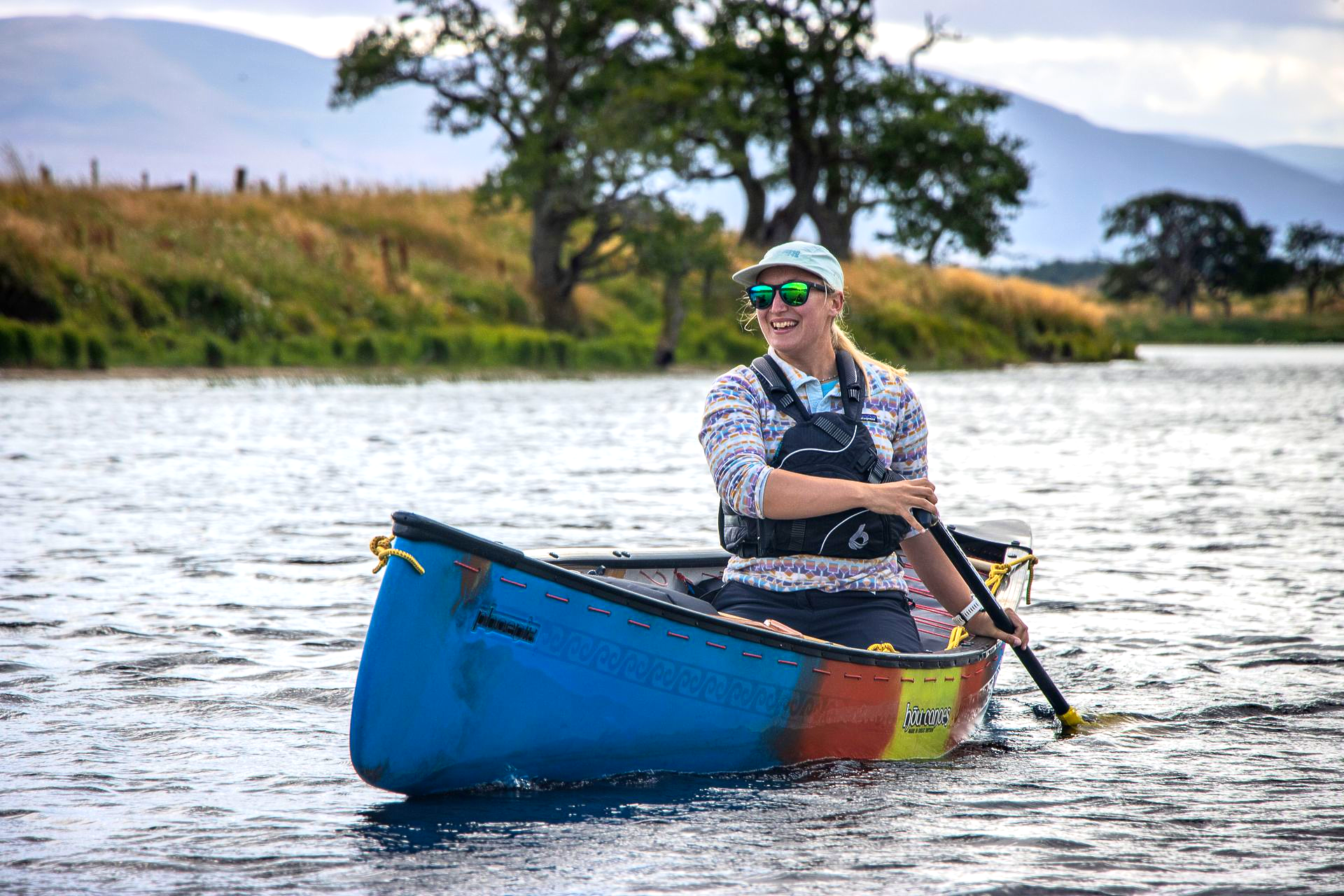 Woman paddles a colorful canoe on a lake, smiling, wearing a hat and life vest.