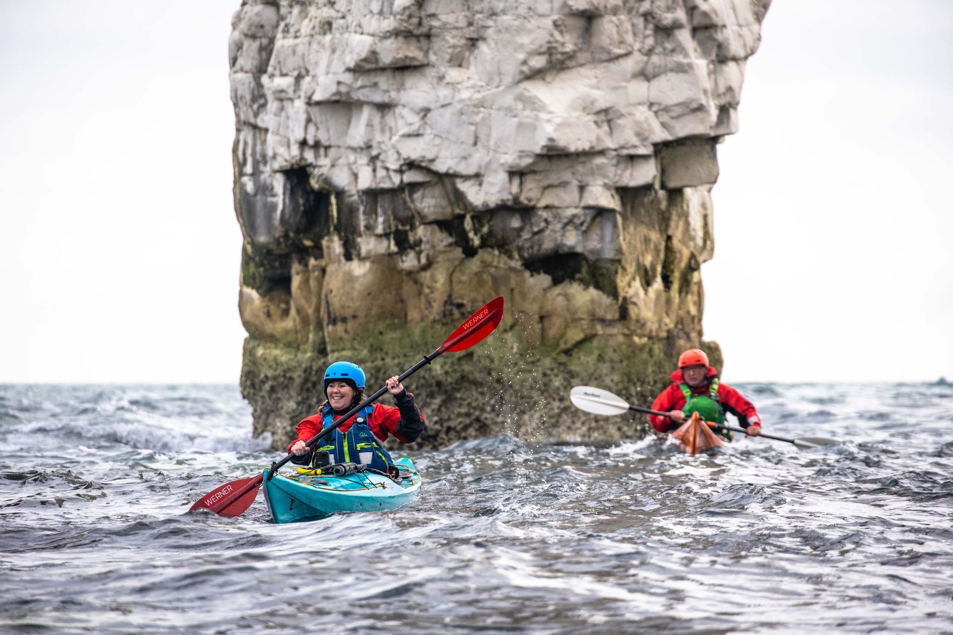 Man kayaking, smiling, with a group in kayaks on a choppy sea near rocky coast under a cloudy sky.