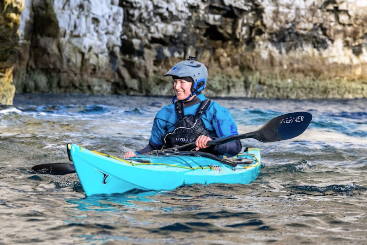 Man in yellow kayak smiles, paddling with group on water near rocky coast under cloudy sky.