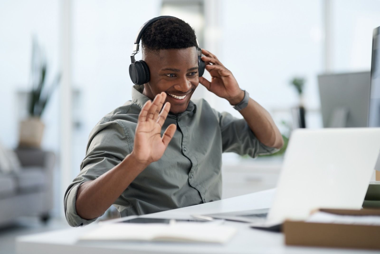 Man with headphones waving at a laptop during a video call in an office.