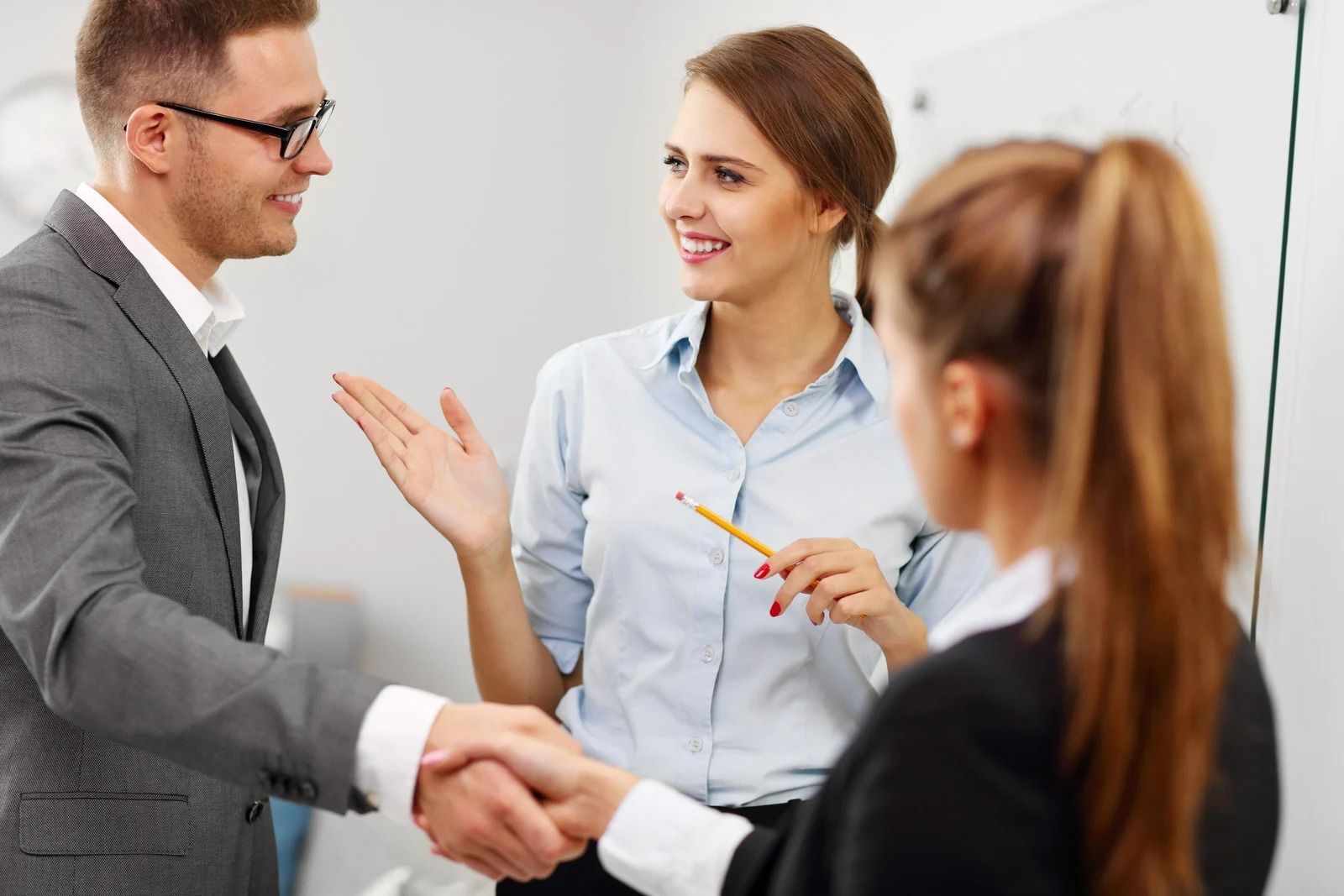 Three people in business attire; man shaking hands with a woman near a whiteboard.
