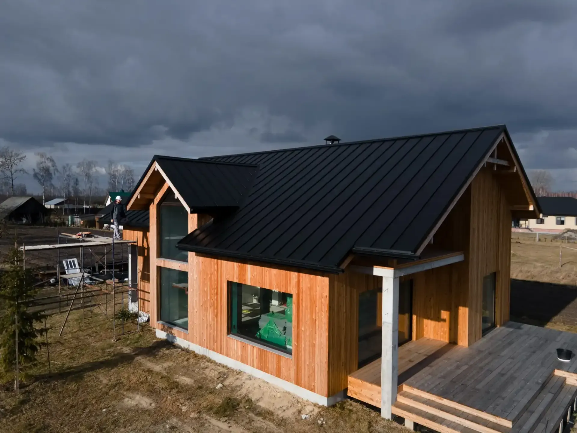 Wooden house with black roof, large windows, and porch under a cloudy sky.
