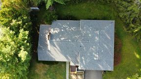Overhead view of a house with a gray roof, surrounded by green trees and grass, and a black driveway.