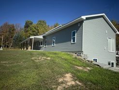 Gray-sided house on a grassy hill under a blue sky, surrounded by trees.