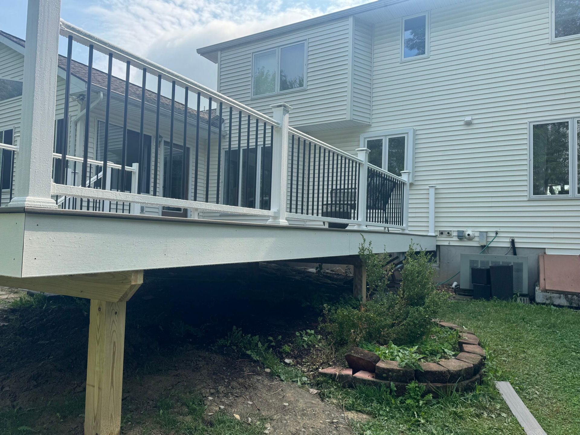 Back deck with white railings, black balusters, and a two-story house with white siding and a small garden.