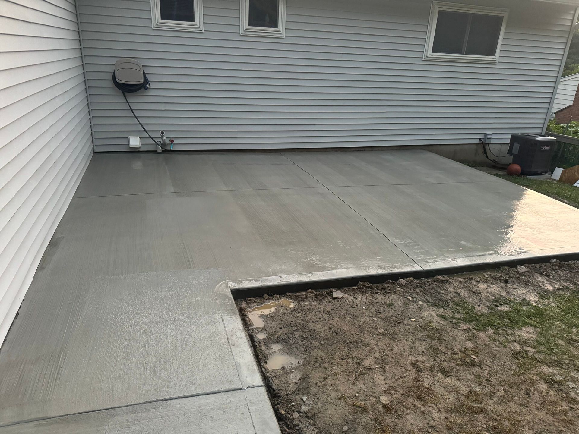Newly poured gray concrete patio next to a house with white siding and small windows.