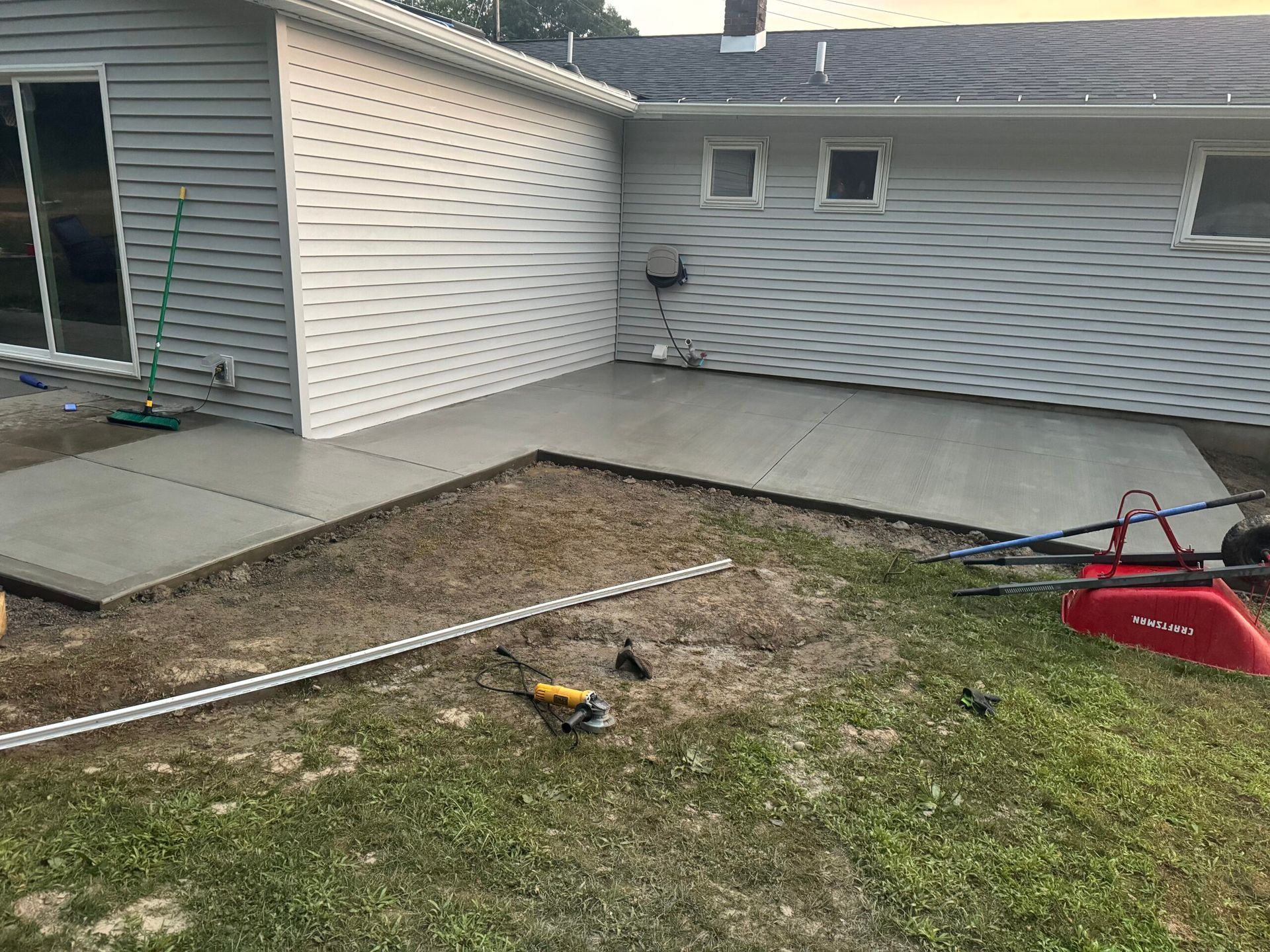 Newly poured concrete patio beside a house with sliding glass door. A red wheelbarrow and tools sit nearby.