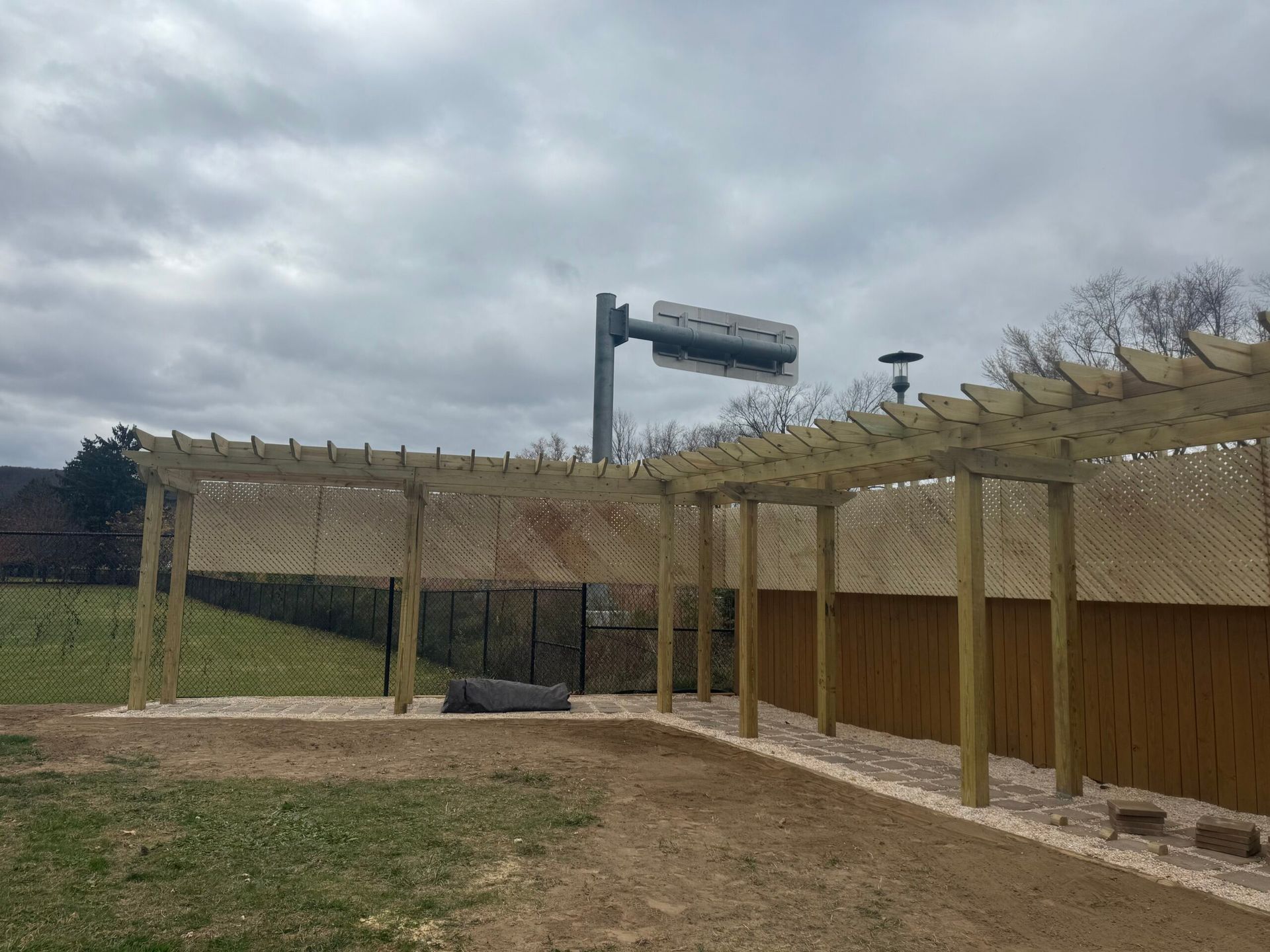 Wooden pergola with gravel path next to a brown fence and a green lawn under a cloudy sky.