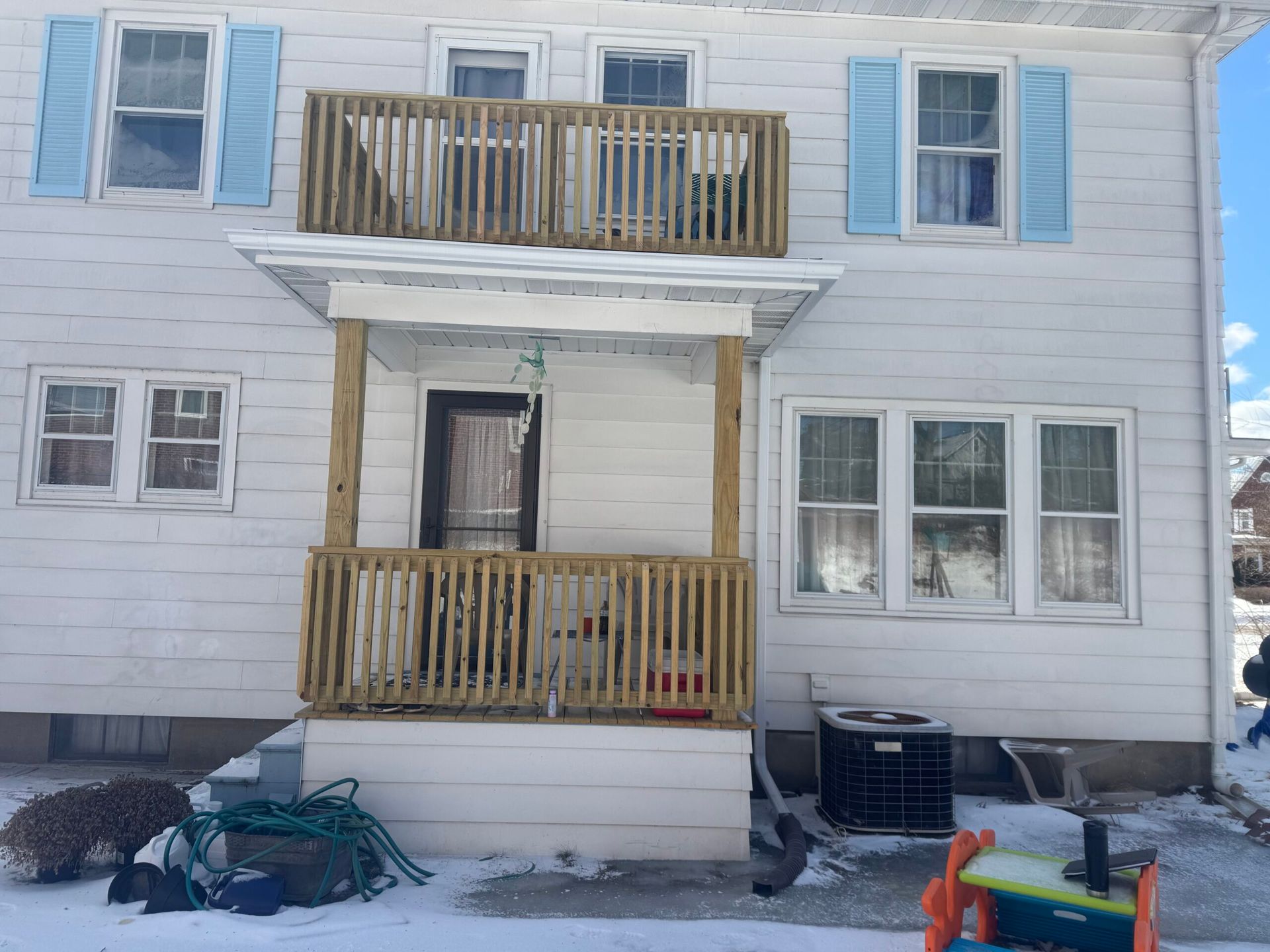 Two-story white house with light blue shutters. A wooden deck and porch are visible. Snow covers the ground.