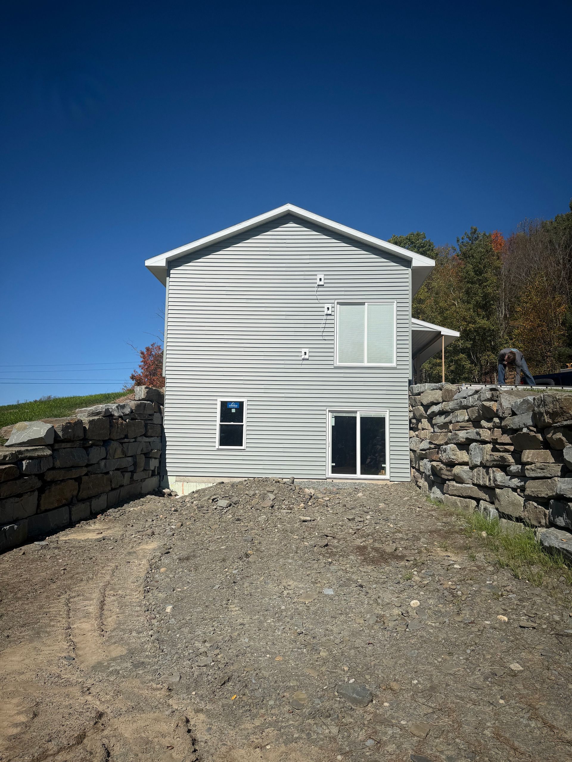 Gray two-story house with horizontal siding, small window, and sliding glass door. A stone wall surrounds the front.
