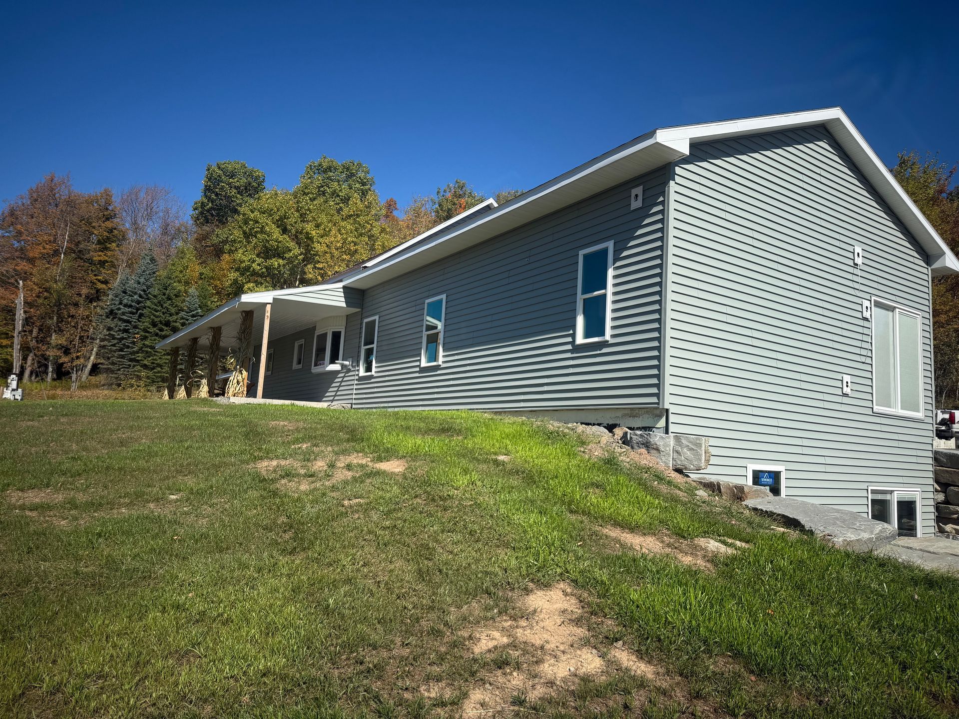 Gray-sided house with white trim, sitting on a grassy hill under a blue sky, surrounded by trees.