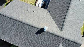 Gray asphalt shingle roof on a house, angled view. Includes chimney, vents, and slight vegetation.
