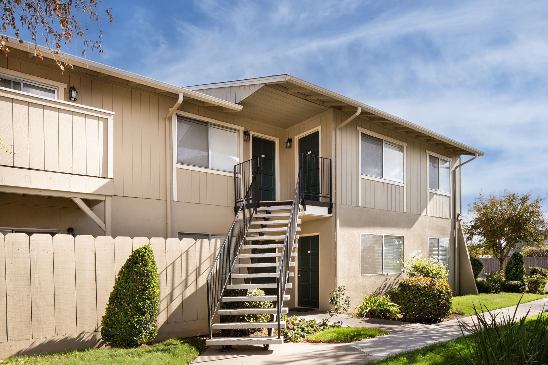 Two-story tan apartment building with exterior stairwell, black doors, and green landscaping.