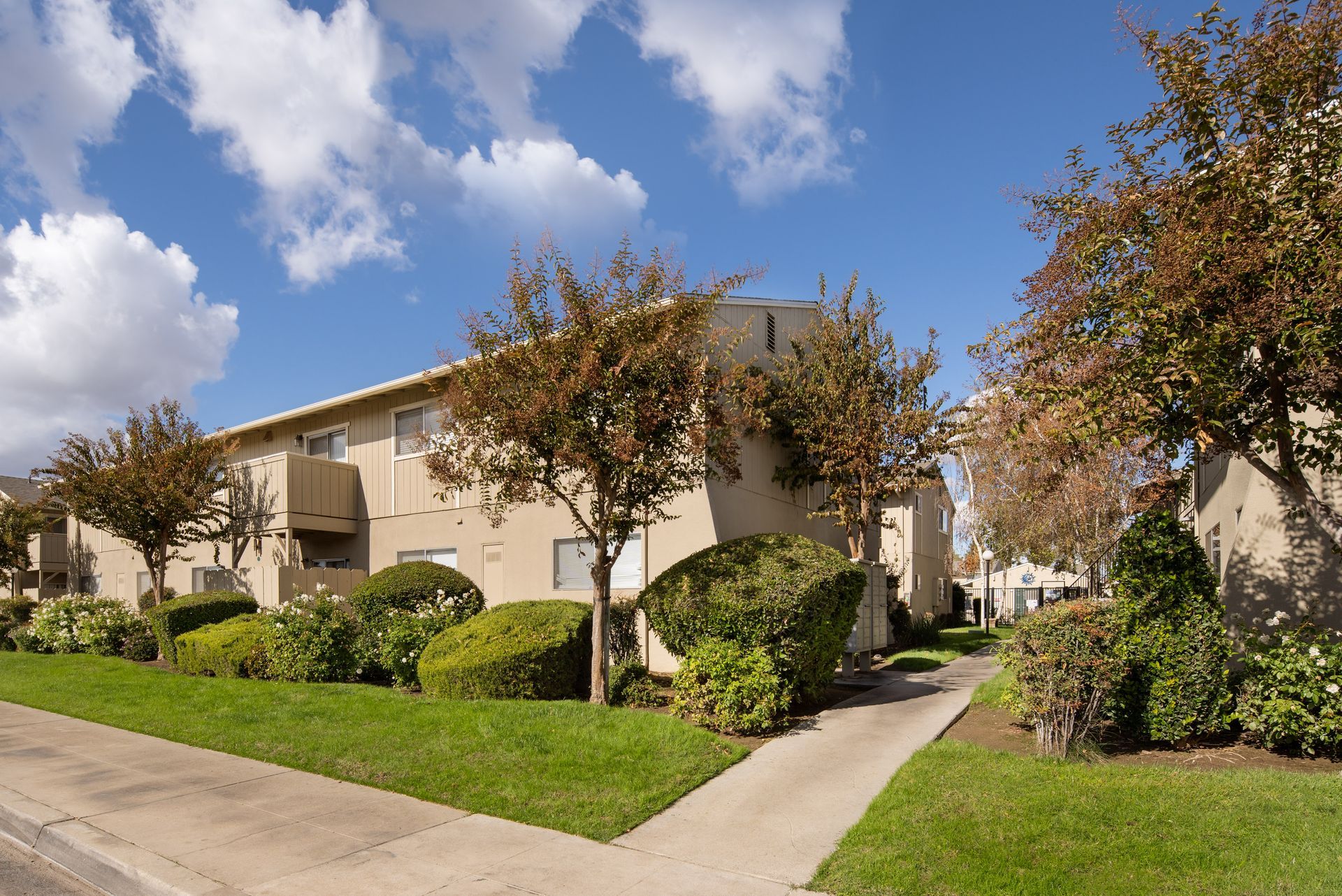 Two-story beige apartment building with manicured landscaping and a sidewalk under a blue sky.