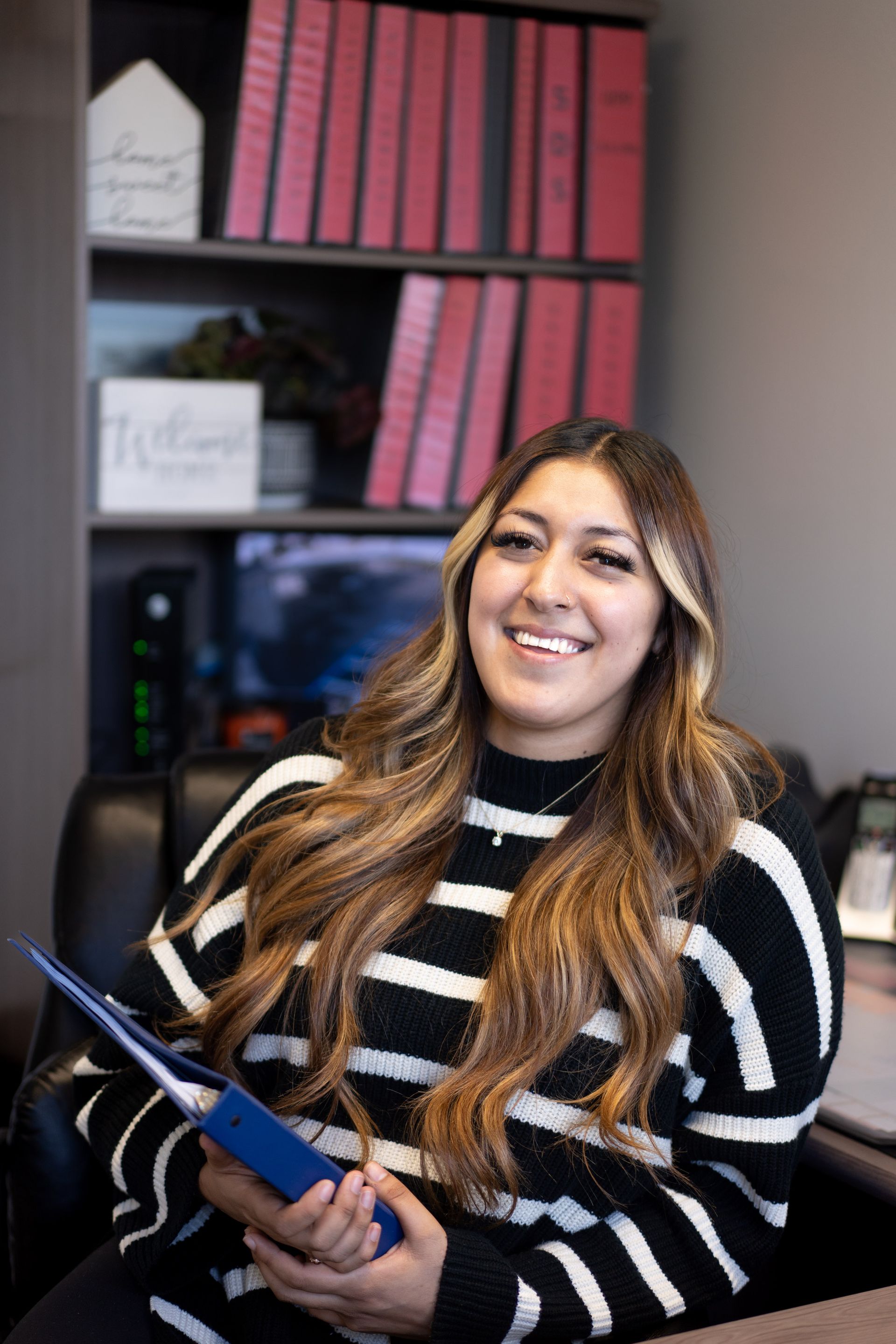 Woman smiling, holding blue folder, in office setting. Wearing black/white striped sweater, with binders on shelves.