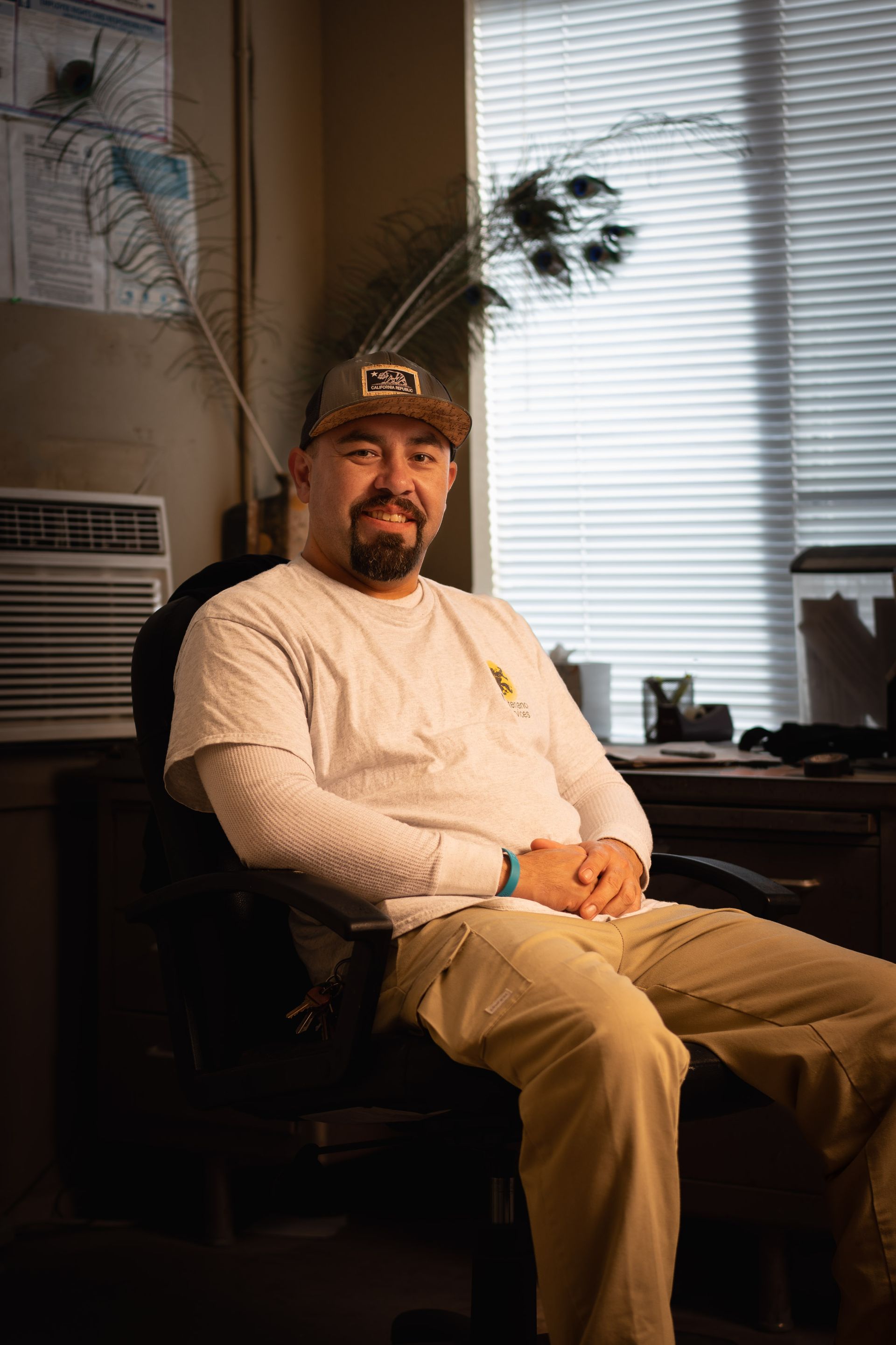 Man wearing cap and casual clothes, sitting at desk, smiling. Peacock feather behind him.