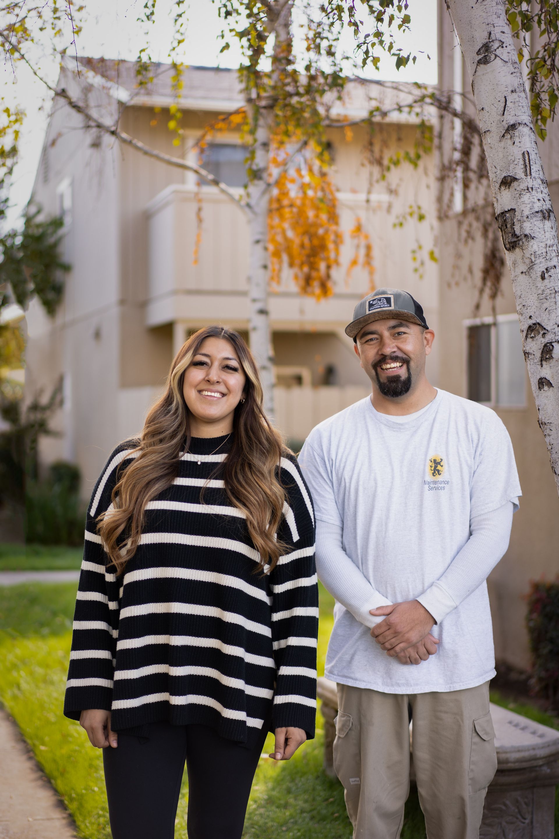 Woman and man standing in front of apartments. Woman in striped sweater, man in a hat.