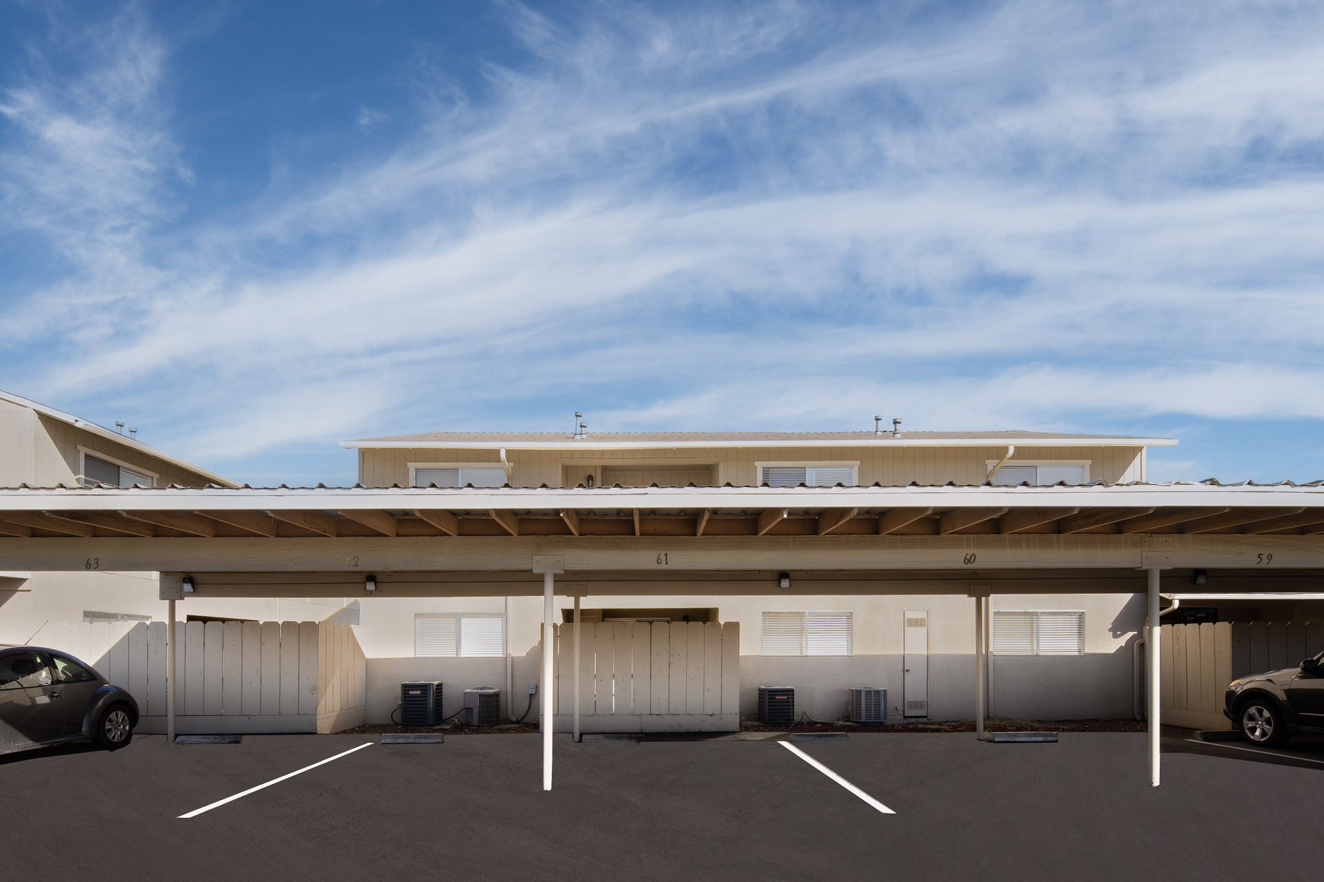 Carport with parked cars under a beige, two-story building against a blue sky with wispy clouds.