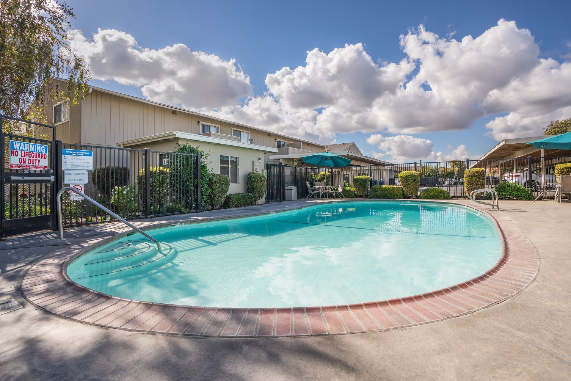 Pool with clear water surrounded by brick and concrete. Buildings and blue sky with clouds in the background.