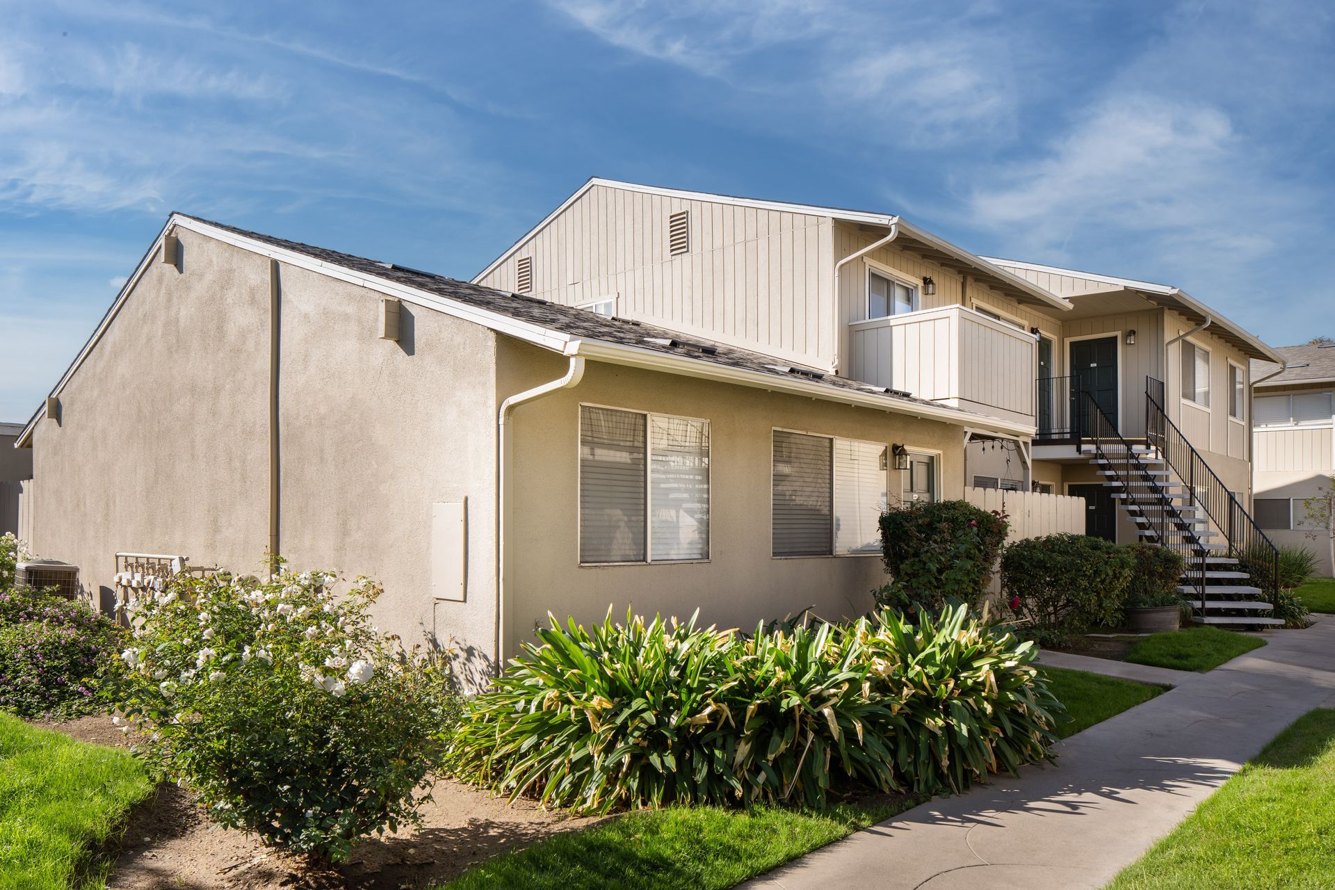Two-story beige apartment building with bushes, a walkway, and a bright blue sky.