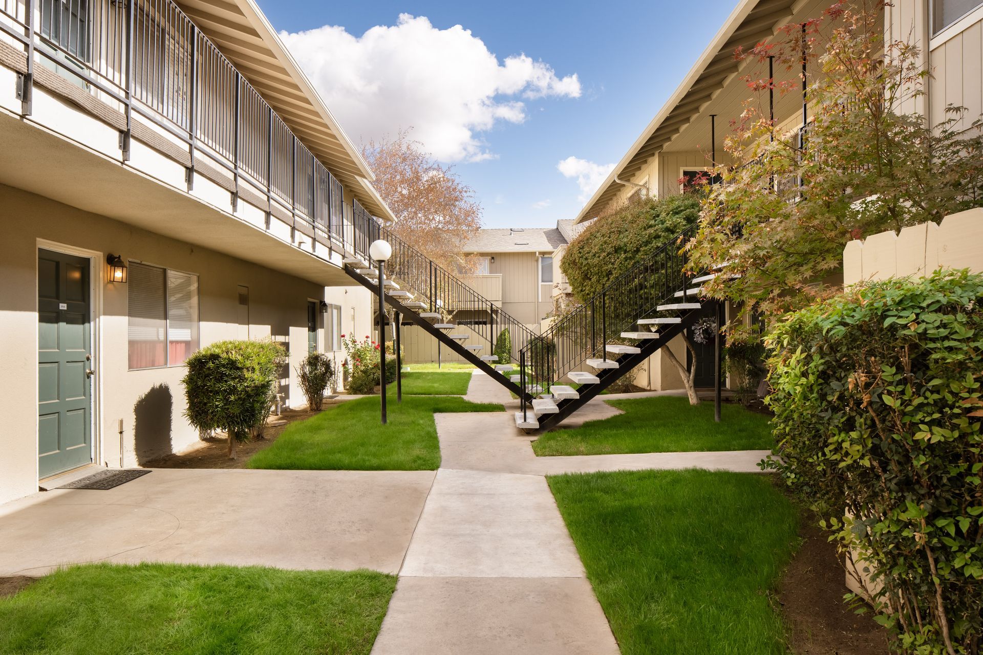 Apartment complex courtyard with walkways, green grass, and staircases leading to upper units. Blue sky overhead.