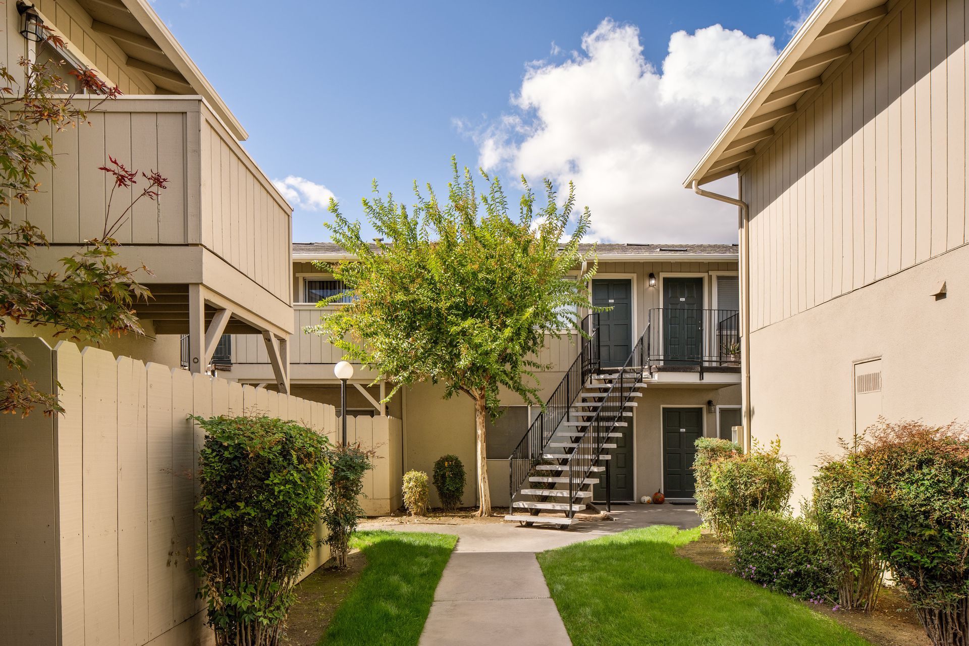 Courtyard of a tan apartment complex with a tree, bushes, and a blue sky.