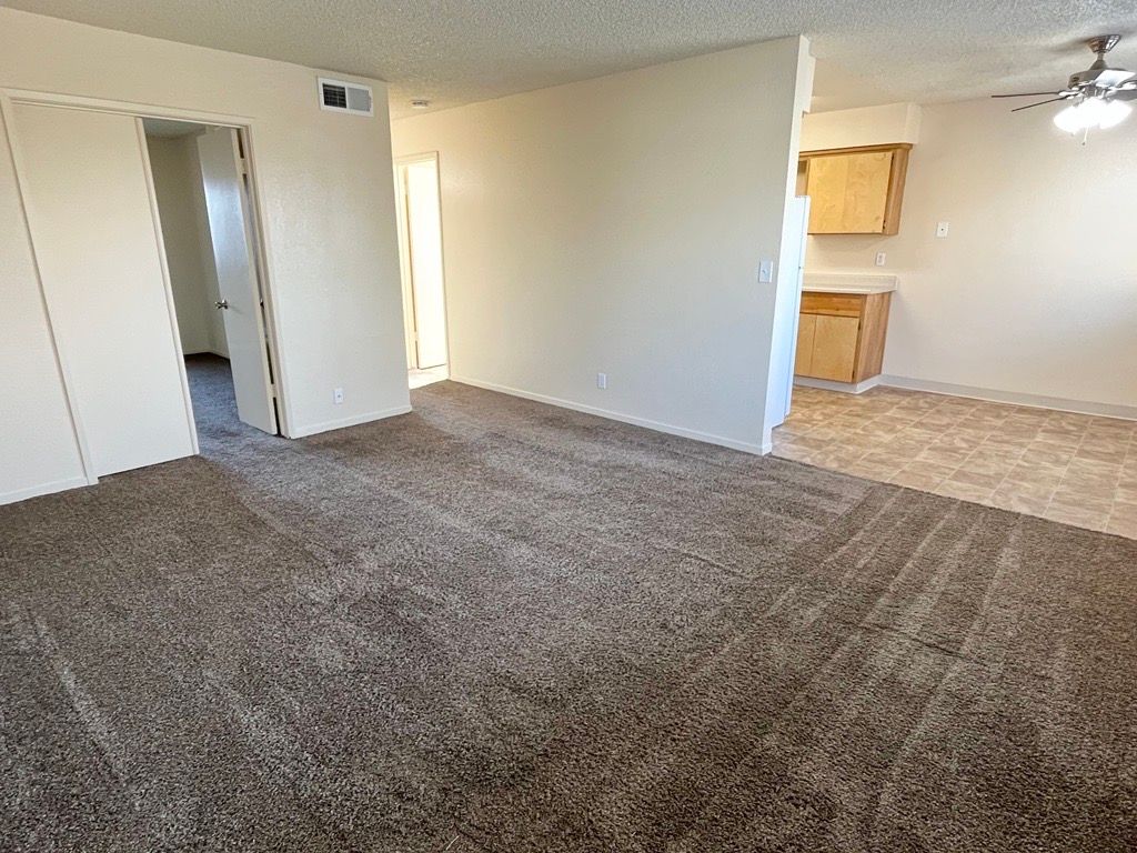 Empty living room with brown carpet, beige walls, and kitchen in the background.